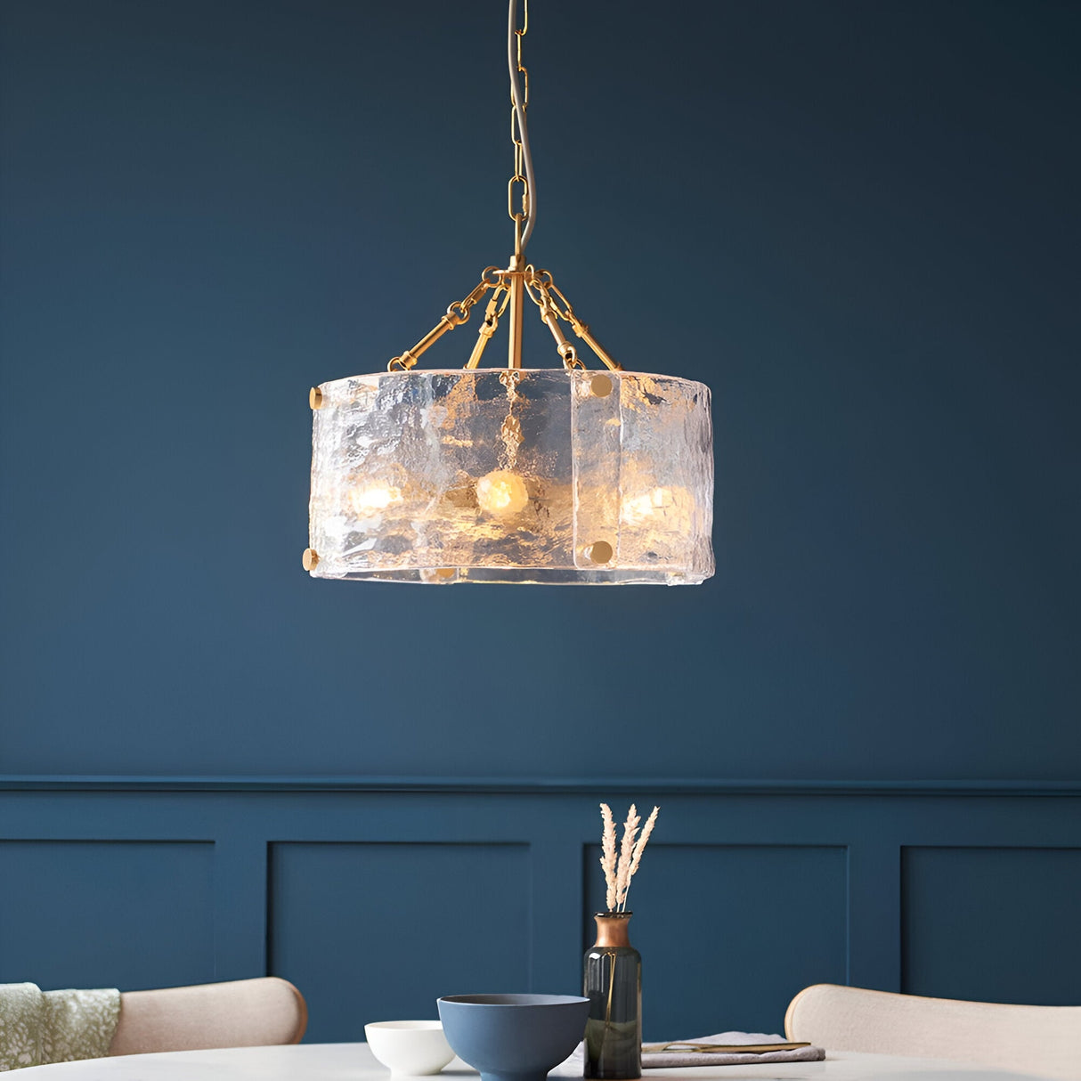 Brockmont 4-Light Pendant in brass with a cylindrical drum shade, textured glass panels, and chain suspension, lit above a dining table.