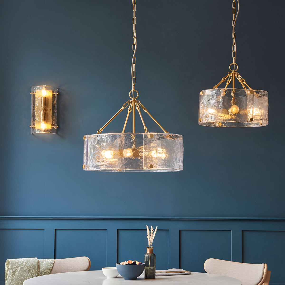 Brockmont 4-Light Pendant in brass with a textured glass drum shade, shown in a dining room with a matching pendant and wall light on a blue panelled wall.