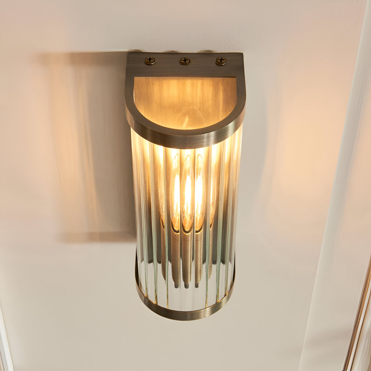 Endon Rothbury wall light in antique brass with ribbed clear glass diffuser and lit E14 bulb, mounted on a pale wall.