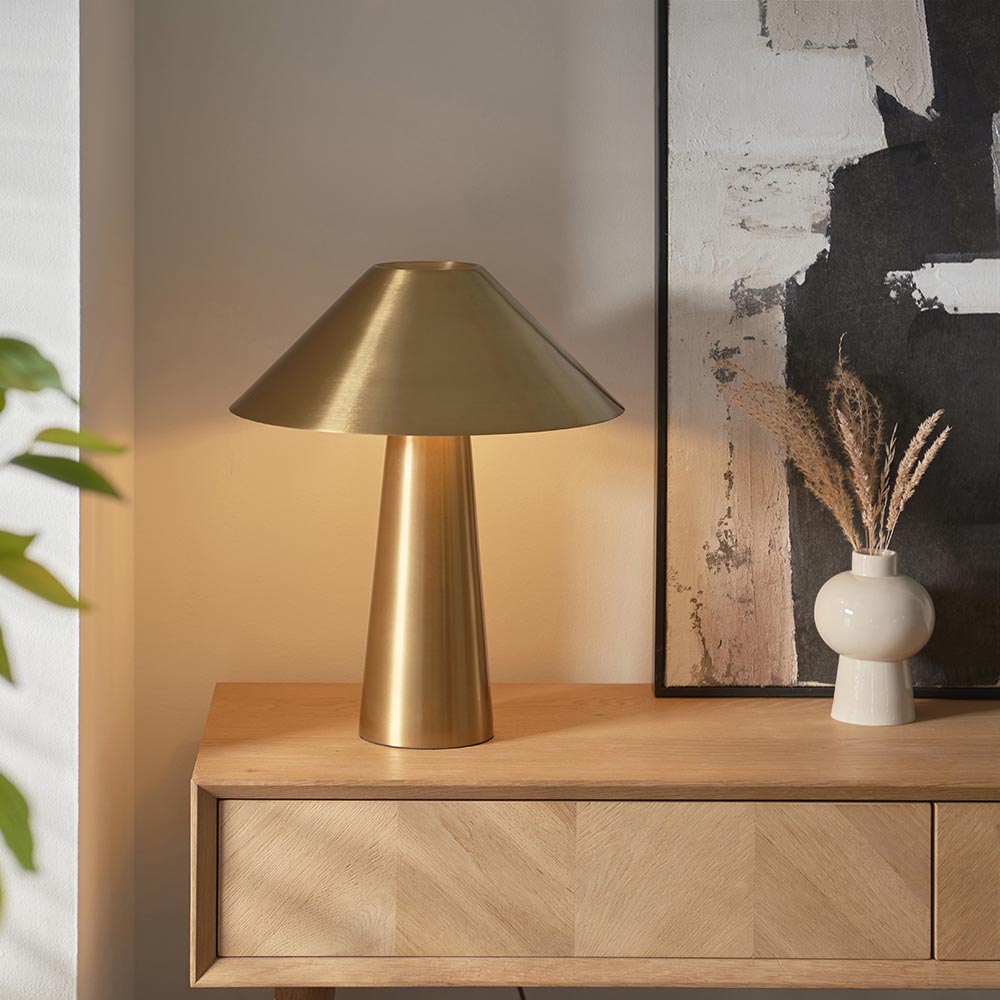 Endon Brockbourne table lamp in brushed antique brass with a wide conical shade and tapered base, styled on an oak sideboard beside a ceramic vase.