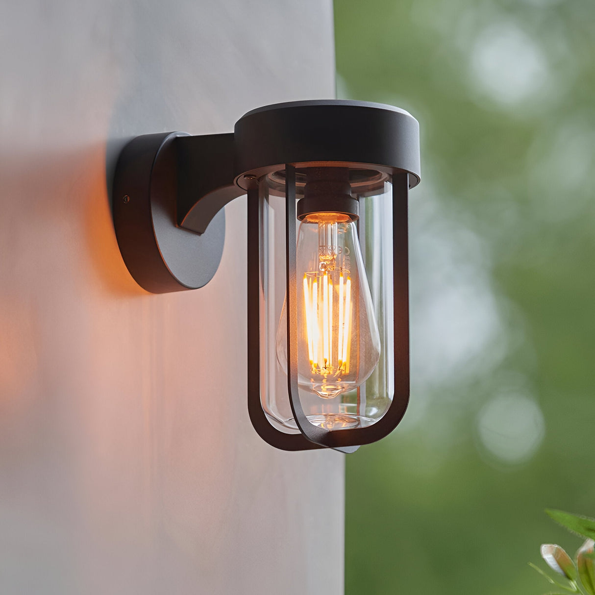 Caldwell Wall Light in matt black die-cast aluminium with a clear glass shade and visible E27 bulb, mounted outdoors against a light wall with greenery beyond.