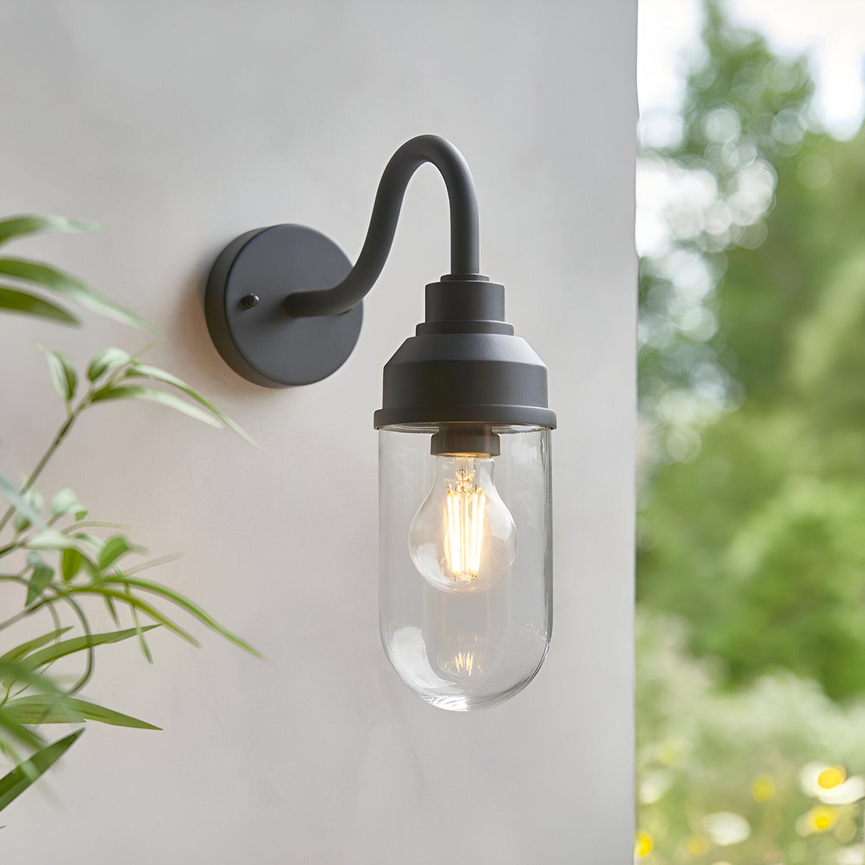 Endon Cranworth matt black wall light with curved arm and clear glass shade housing an E27 filament bulb, mounted on a pale exterior wall beside foliage.