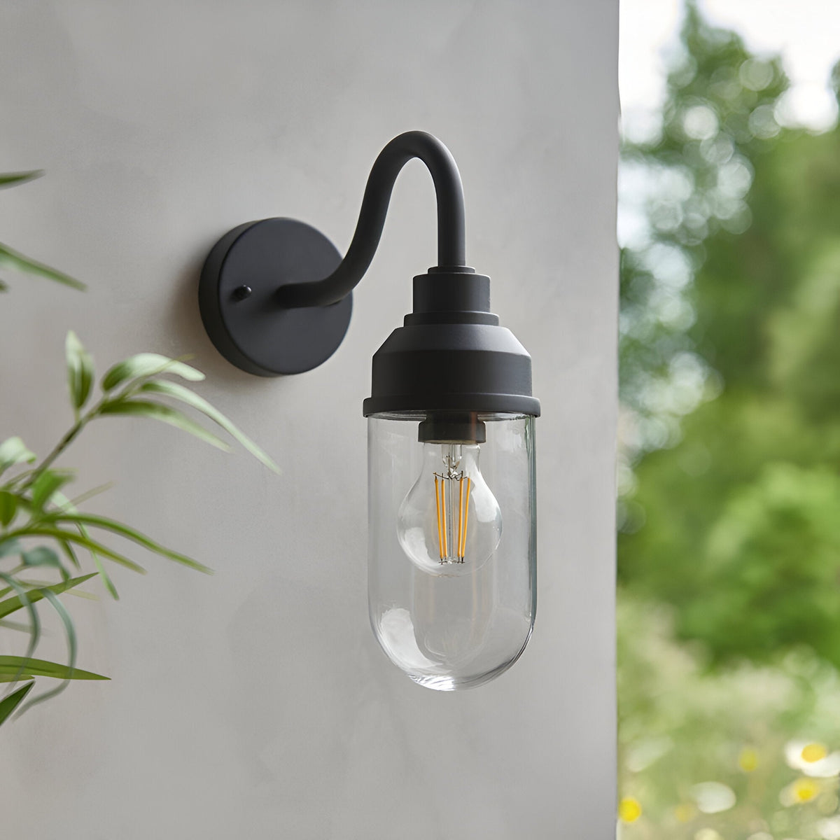 Endon Cranworth curved-arm wall light in matt black aluminium with a clear glass shade and E27 filament bulb, mounted on a light exterior wall.