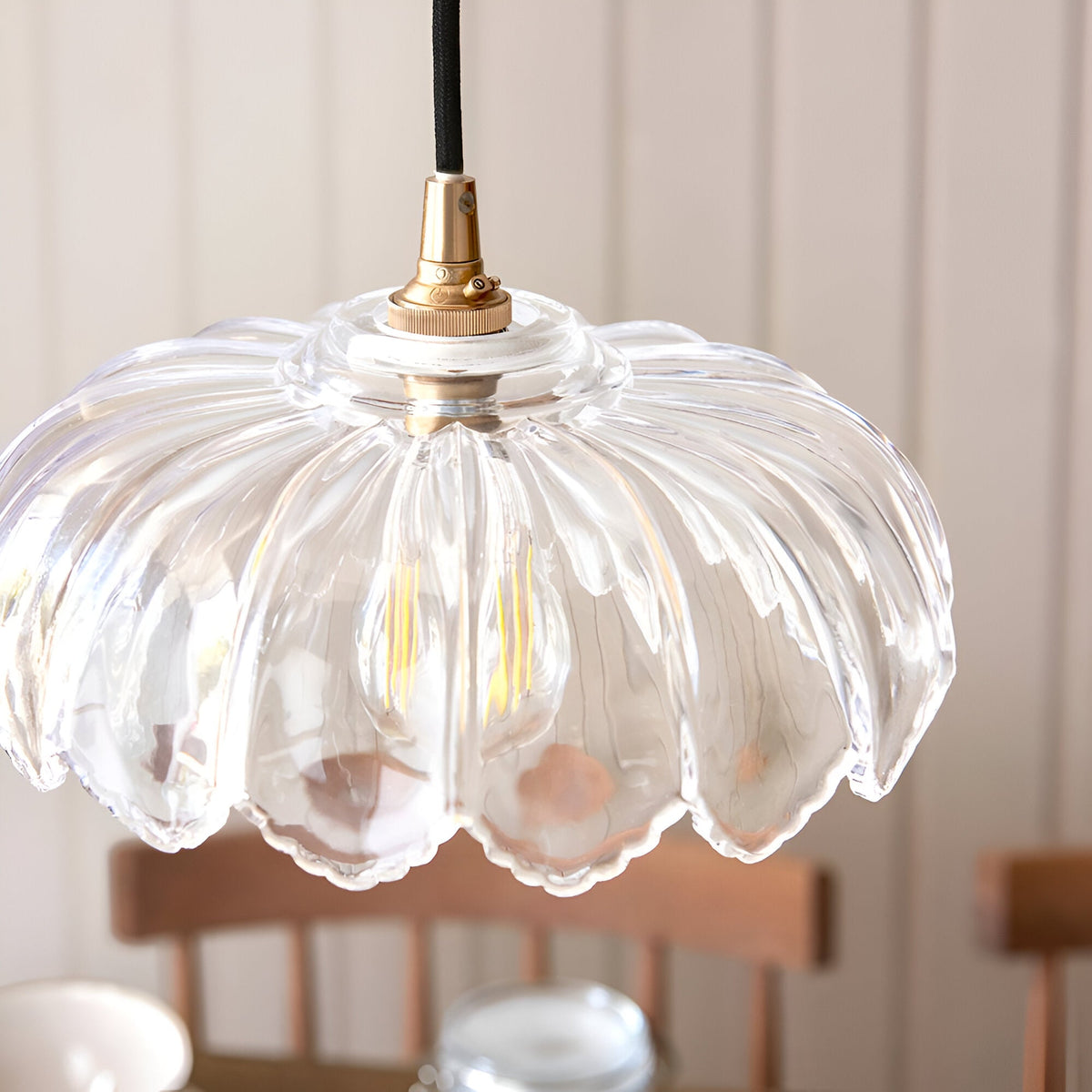 Ashwick clear glass pendant shade with scalloped edge and brass fitting, shown close-up with visible bulb, in a room with wooden chairs.