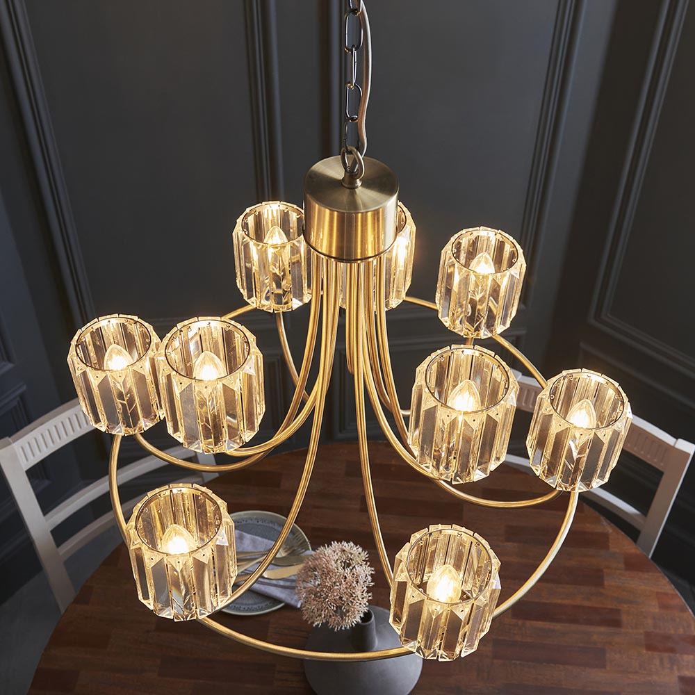 Clementine 9 Light Tiered Chandelier in antique brass with faceted glass shades, shown lit above a dining table in a dark-panelled room.