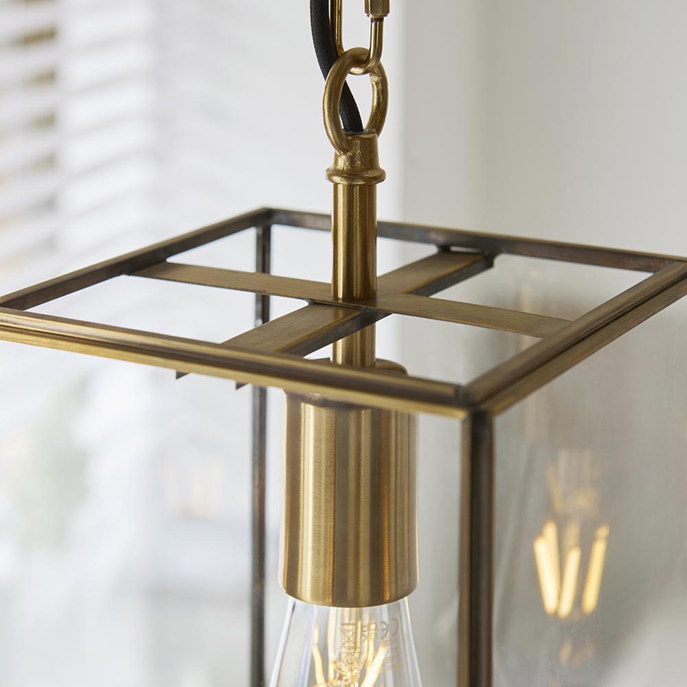 Close-up of the Haddon Pendant in brushed brass, showing the lantern-style frame, clear glass panel, E27 bulb holder, and fabric flex with chain link.