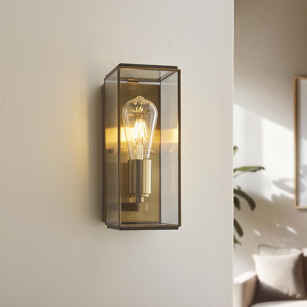 Haddon Wall Light in antique brass, a box-lantern fixture with clear glass panels and exposed filament bulb, shown mounted on a wall in a living room setting.