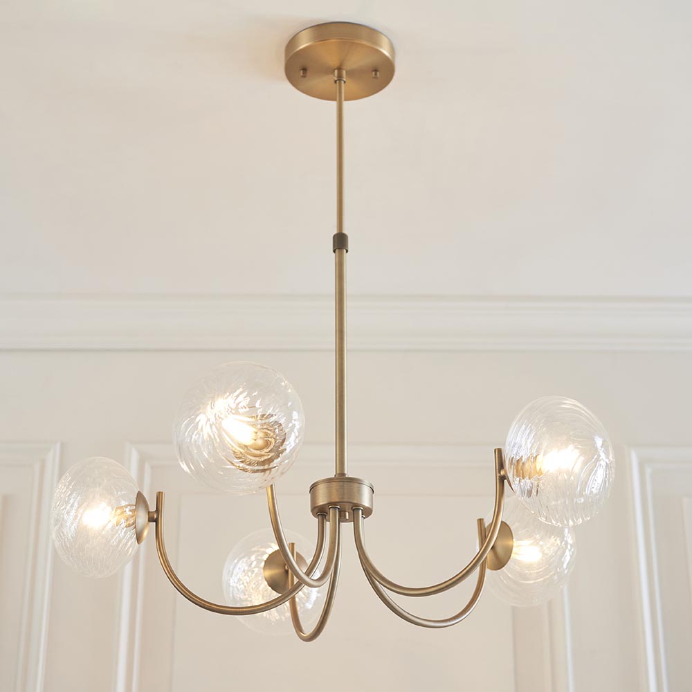 Bramwick 5 Light Pendant in antique brass with curved arms and clear twisted glass globes, shown suspended from a panelled ceiling on an adjustable stem.