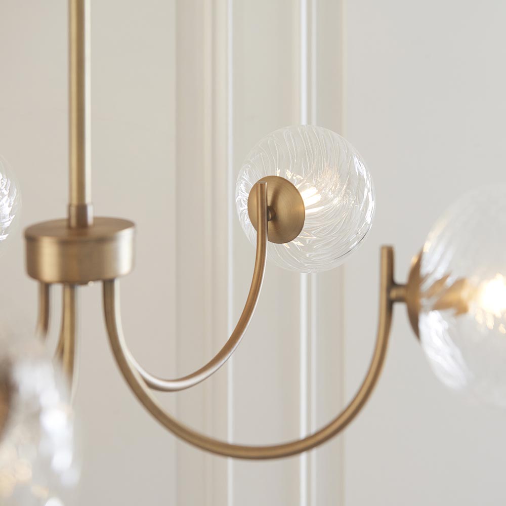 Close-up of the Bramwick 5 Light Pendant in antique brass, showing curved arms and clear twisted glass globe shades against a light interior wall.