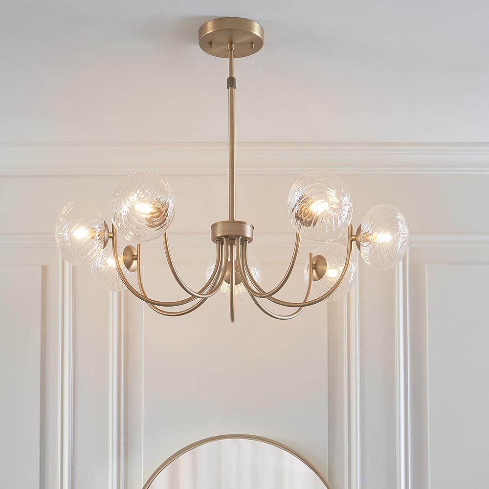 Endon Bramwick 7 light pendant in antique brass with curved arms and clear twisted glass globes, shown below a ceiling above a round mirror.