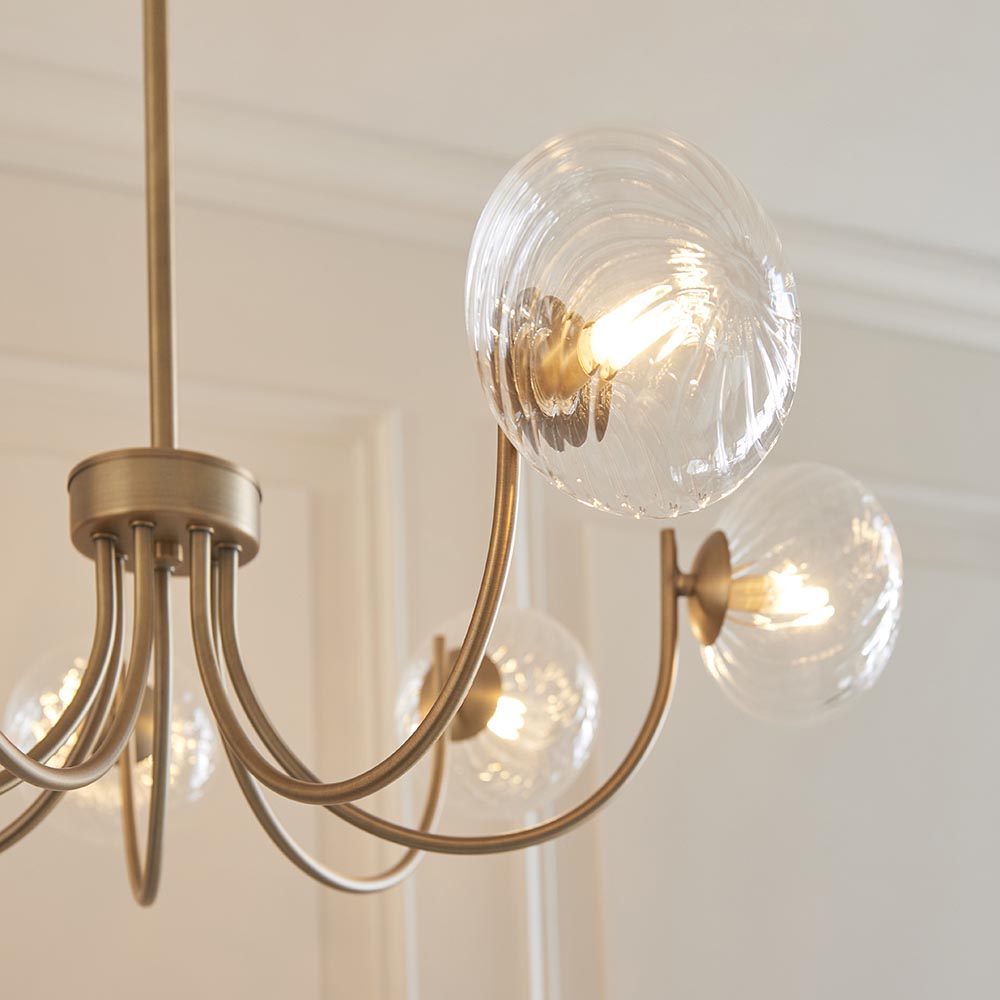 Endon Bramwick 7 Light Pendant in antique brass with curved arms and clear twisted glass globes, shown lit against a panelled cream wall.