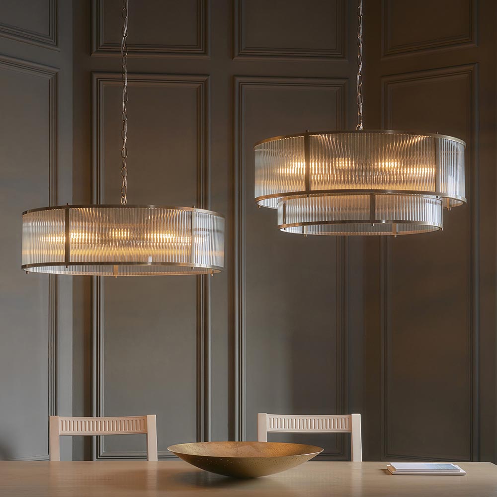 Two Endon Rothbury 8 Light Pendants in antique brass with ribbed glass panel shades, hung on chains above a dining table against a panelled grey wall.