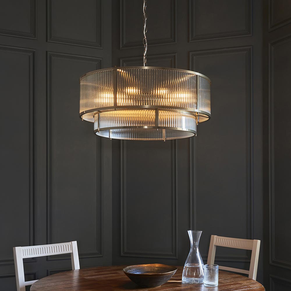 Endon Rothbury 8 light tiered chandelier in antique brass with clear ribbed glass panels, hung above a wooden dining table against dark panelled walls.