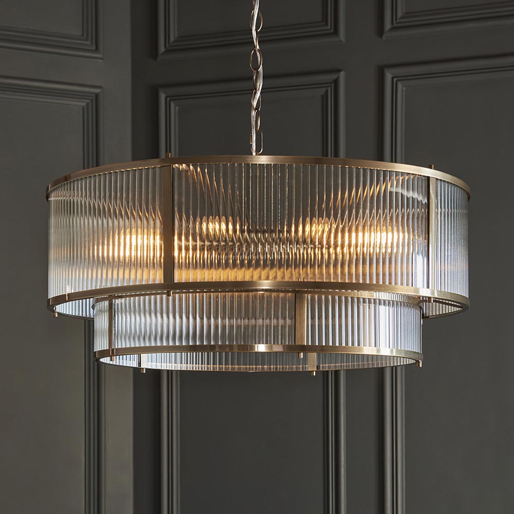 Endon Rothbury 8 light tiered chandelier in antique brass with clear ribbed glass panels over two tiers, suspended from a chain against a grey panelled wall.