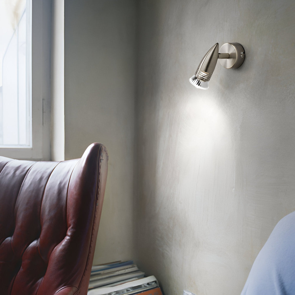 An elegant room with a minimalist design is illuminated by the Omega Single Spotlight in a burnished brass finish, which offers adjustable light direction. The light highlights the beige walls and dark red leather armchair, complemented by natural light streaming through a nearby window. Stacks of books are arranged in the corner, enhancing the sophisticated ambiance.