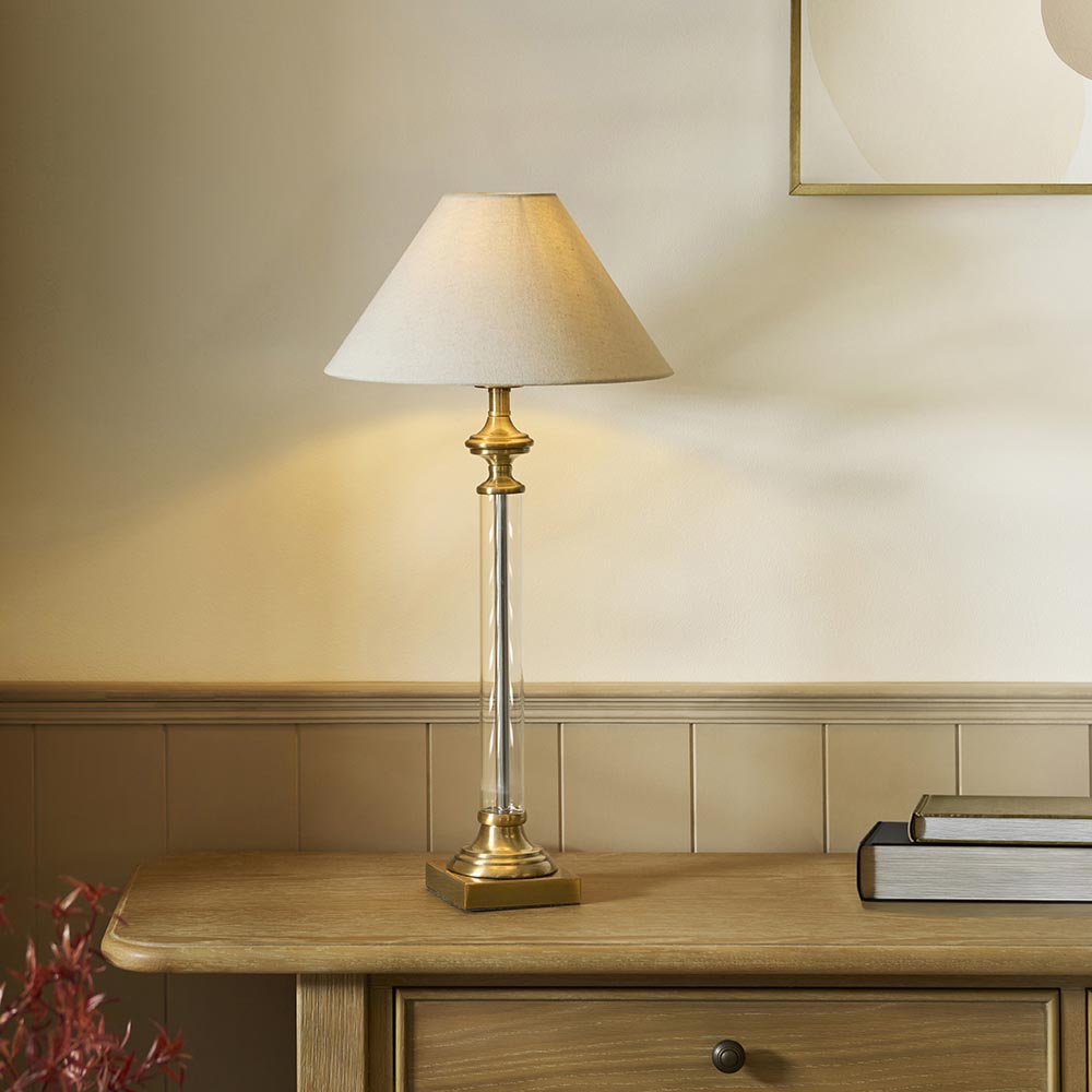 Aldmont Avalon Table Lamp with clear glass column, antique brass base and cap, and tapered natural linen shade, shown on a wooden sideboard.