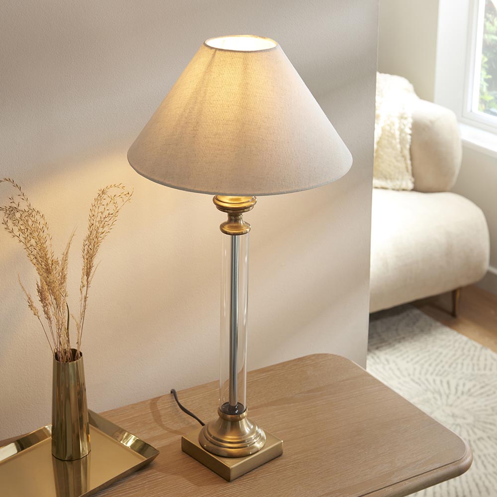 Aldmont Avalon table lamp with clear glass column, antique brass base and cap, and tapered linen shade, lit on a wooden side table.