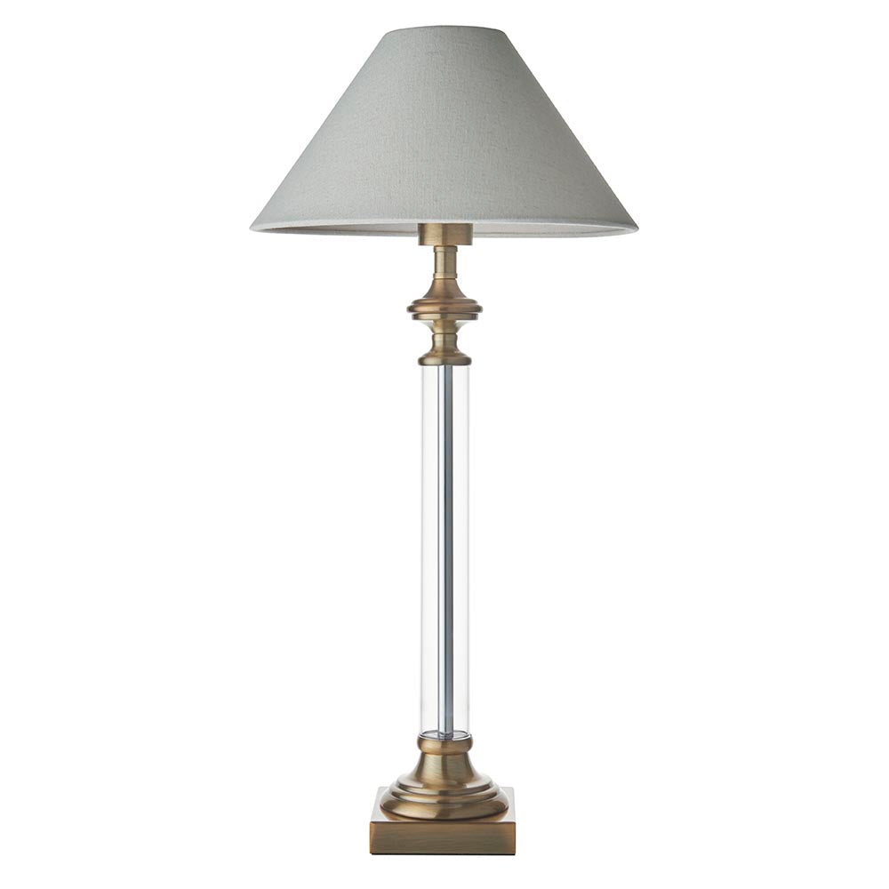Aldmont Avalon table lamp with a clear glass column, antique brass base and cap, and a tapered natural linen shade, shown on a plain white background.