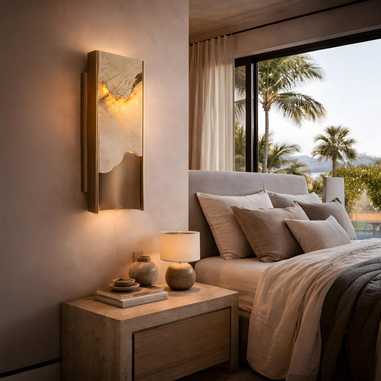A modern bedroom with a big window and palm trees, a neutral bed, a stone nightstand with a lamp and vases, and the Veloris LED Small Carved Alabaster Wall Light in Brushed Brass & Soft White adding warm, marble-like accent lighting.
