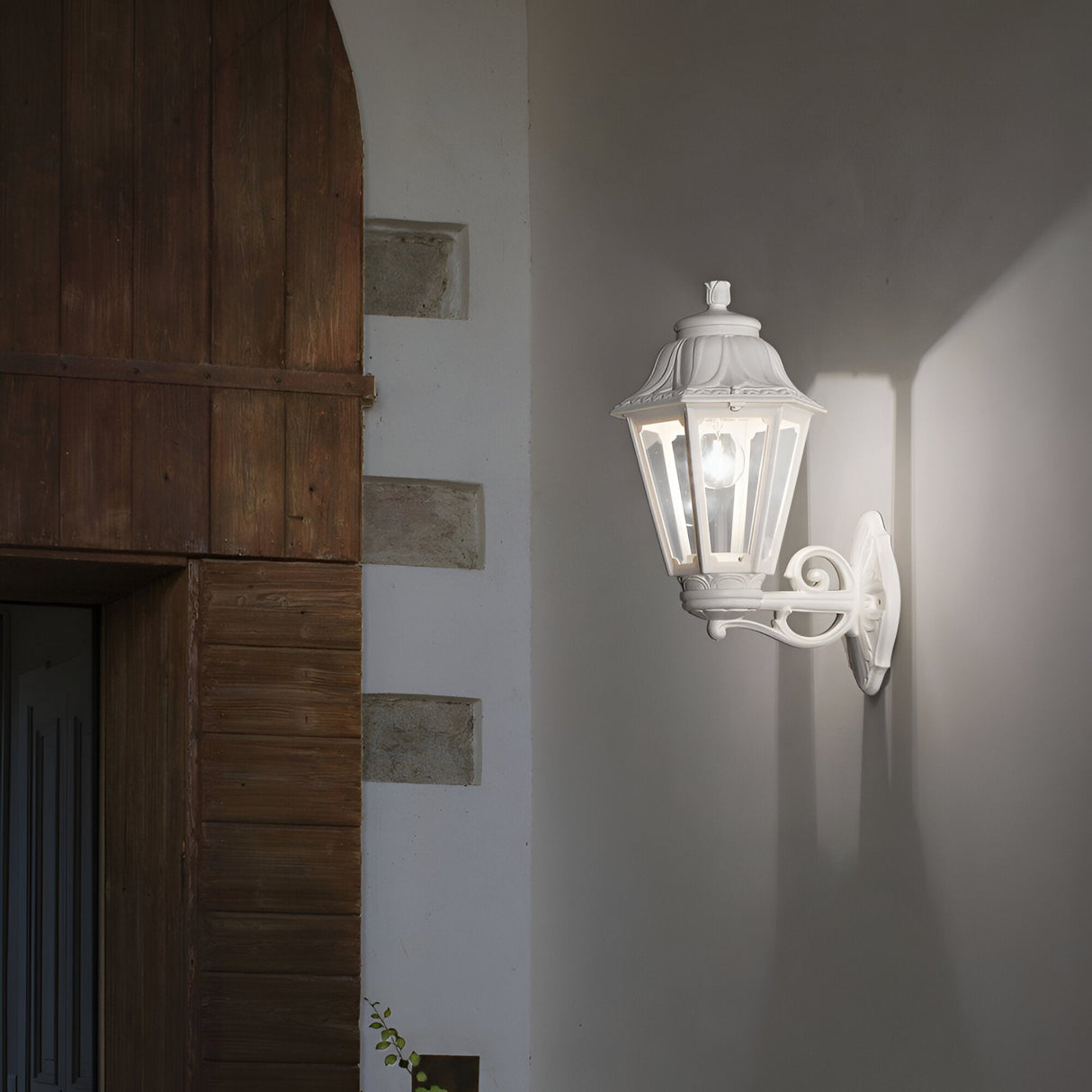 The Gaze Large Outdoor Wall Light - White, with its vintage style and glowing bulb, is elegantly situated on a white wall beside a wooden door. Its design features an anti-yellowing diffuser that projects a warm light and shadow pattern, preserving its charm over time.