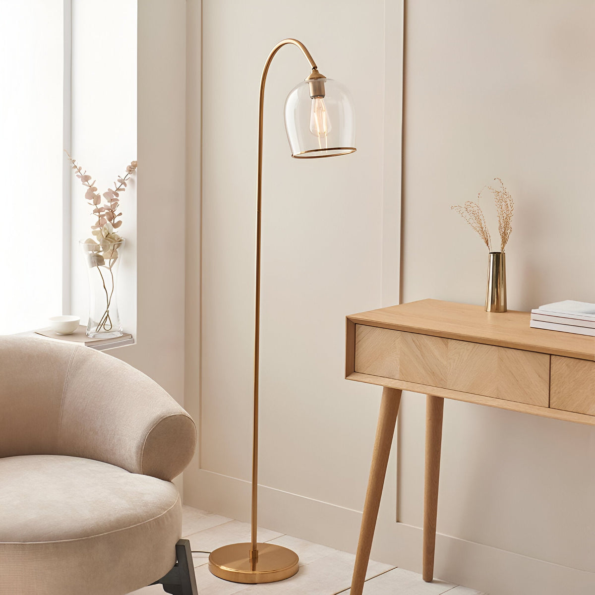Halstead Floor Lamp in aged brass with curved stem and clear domed glass shade, shown beside an armchair and wooden desk in a living room setting.