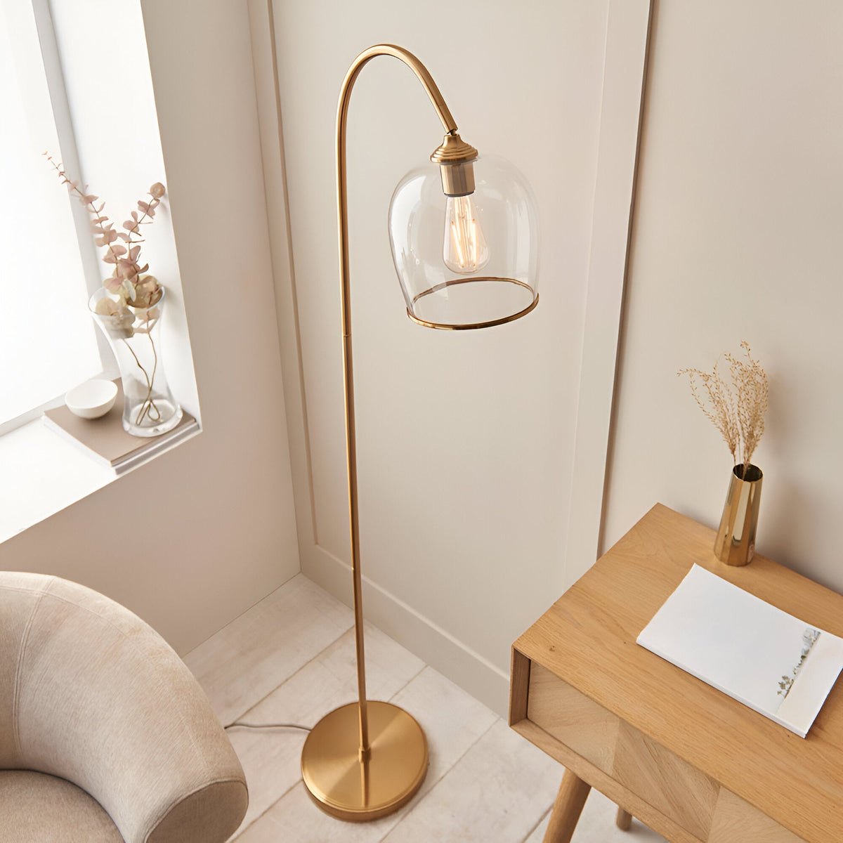Halstead Floor Lamp in aged brass with curved stem and domed clear glass shade, shown beside a wooden side table in a living room setting.