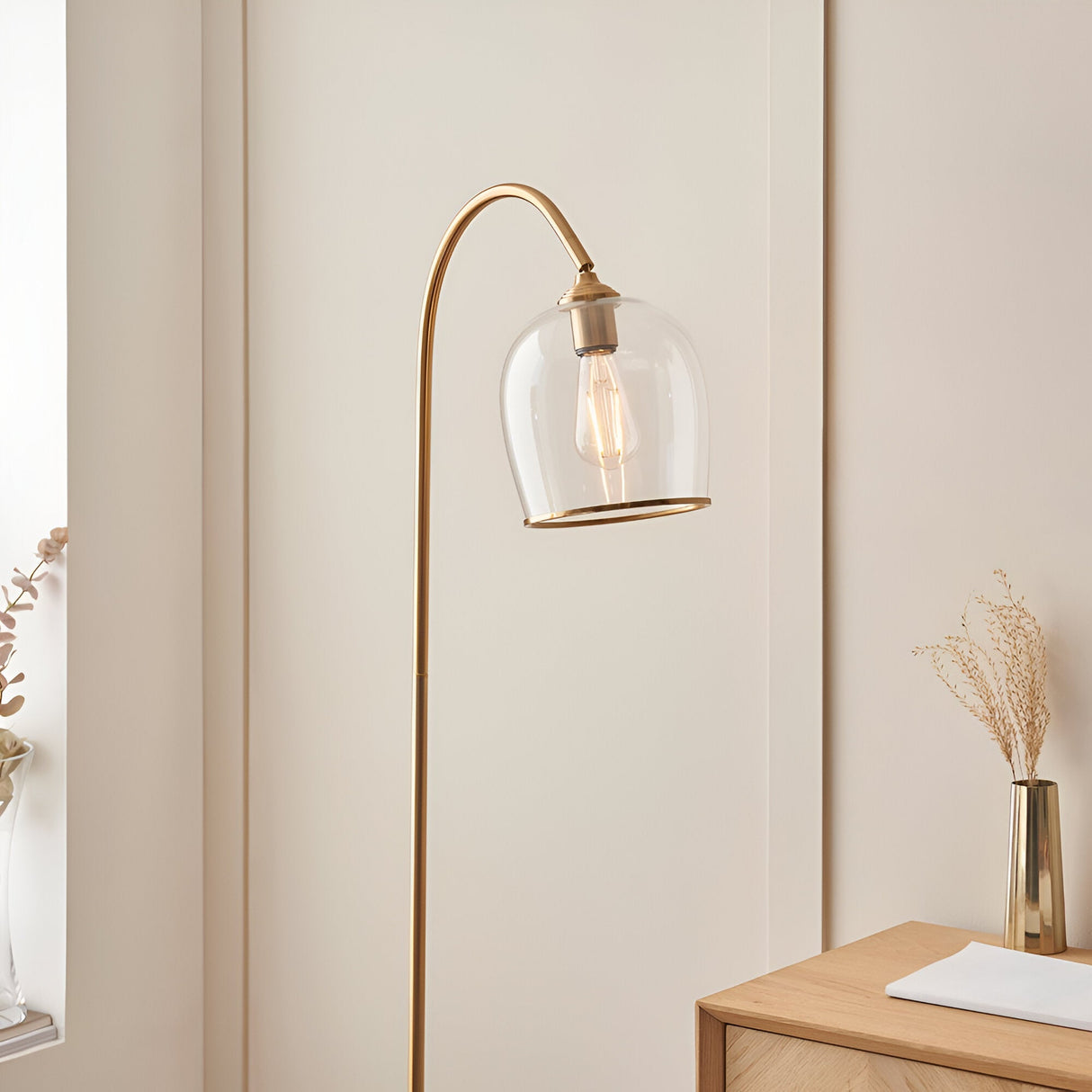 Halstead Floor Lamp in aged brass with a curved stem and domed clear glass shade, shown in a living room beside a wooden cabinet.