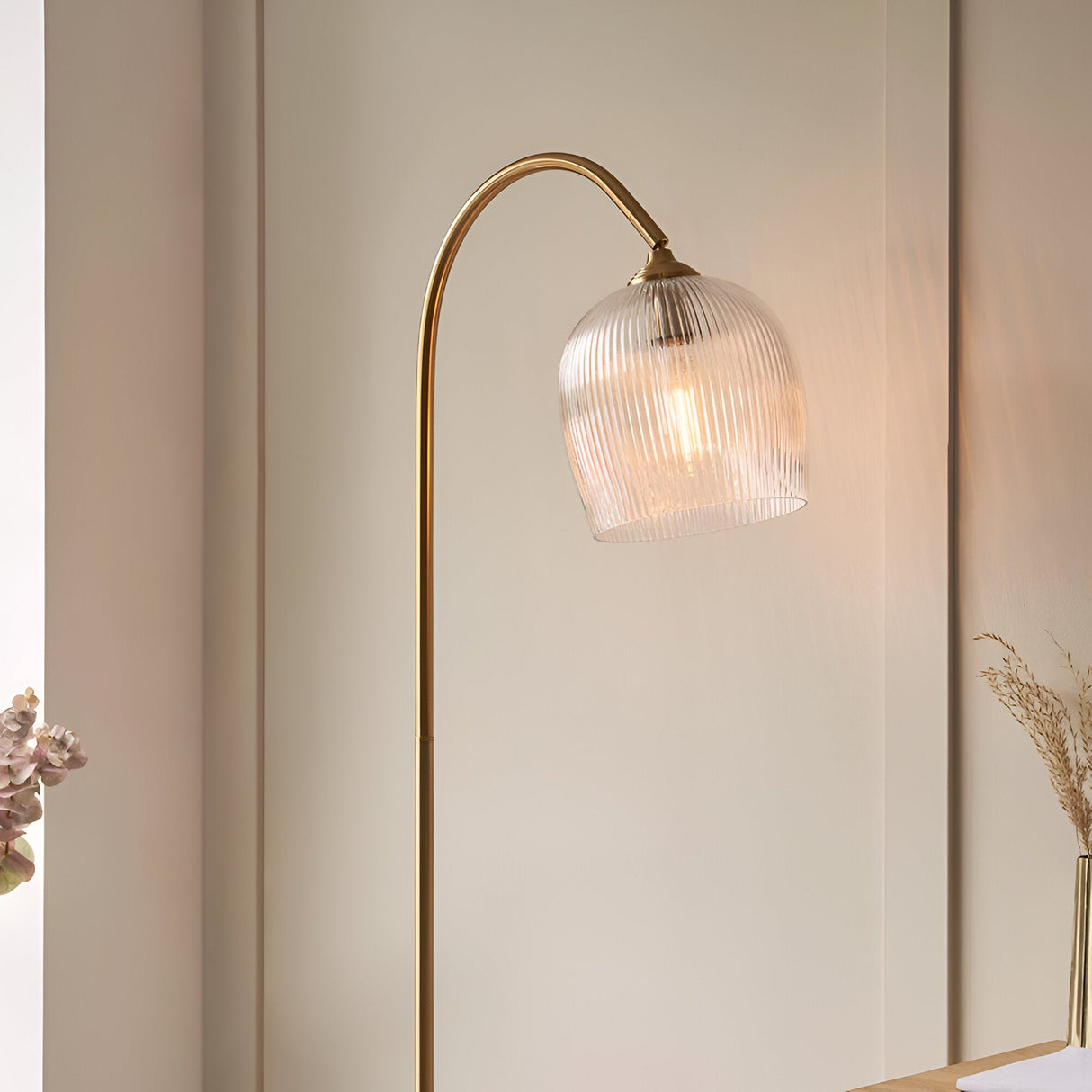 Endon Halstead floor lamp with arched aged brass stem and ribbed clear glass domed shade, lit beside a neutral panelled wall and dried floral stems.