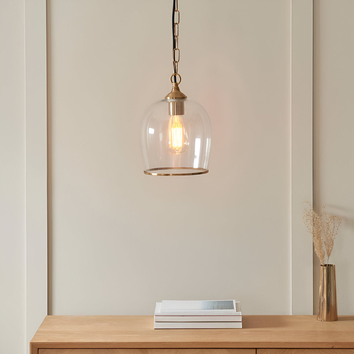 Halstead Pendant in brass, clear glass dome shade with brass edge and chain, hung above a wooden console with books and a vase.