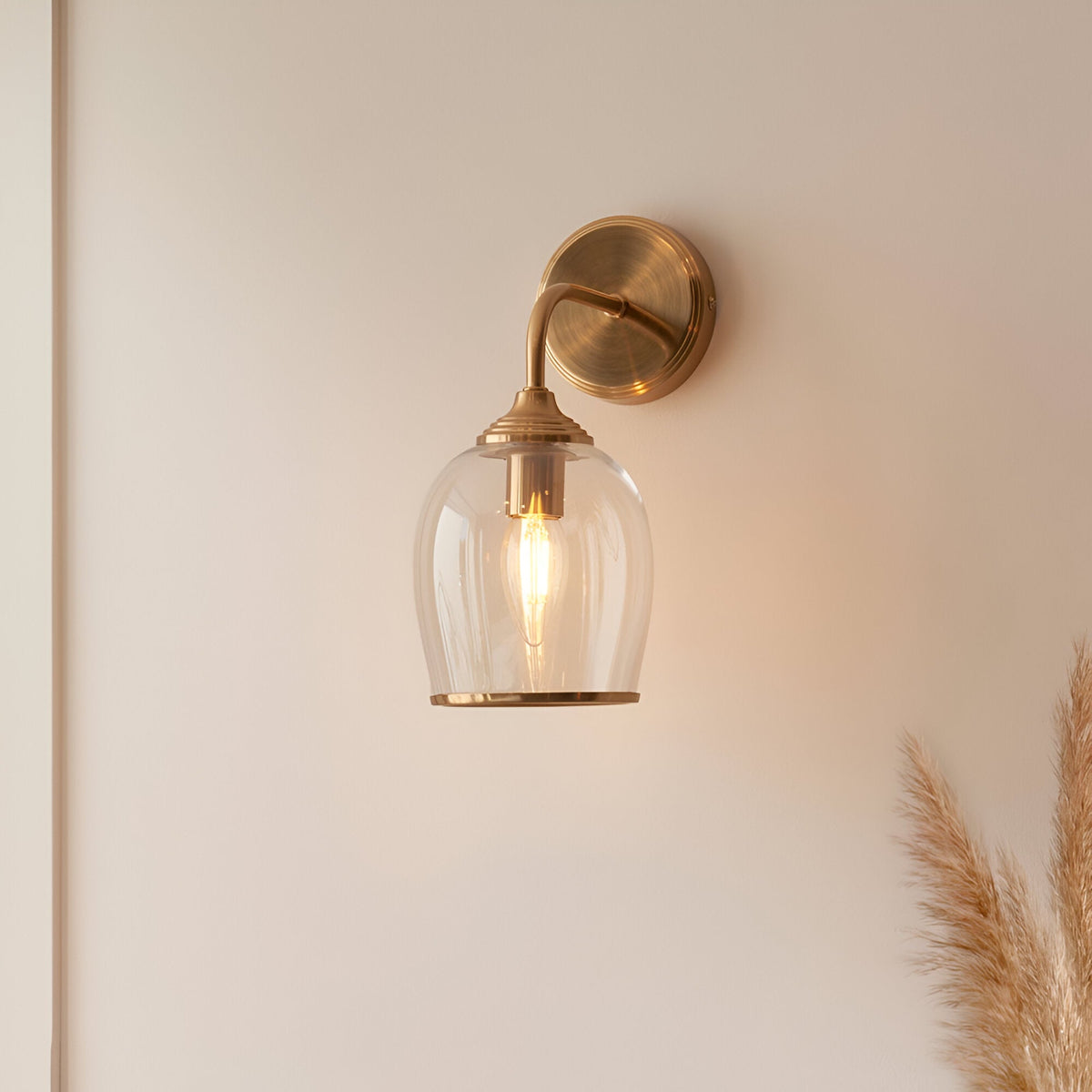 Halstead Wall Light in brass with a clear domed glass shade, curved arm, and round back plate, mounted on a light wall.