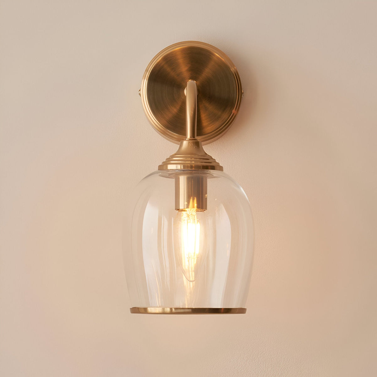 Halstead Wall Light in brass with a curved arm, round back plate, and clear domed glass shade holding a lit E14 bulb, shown on a plain light background.
