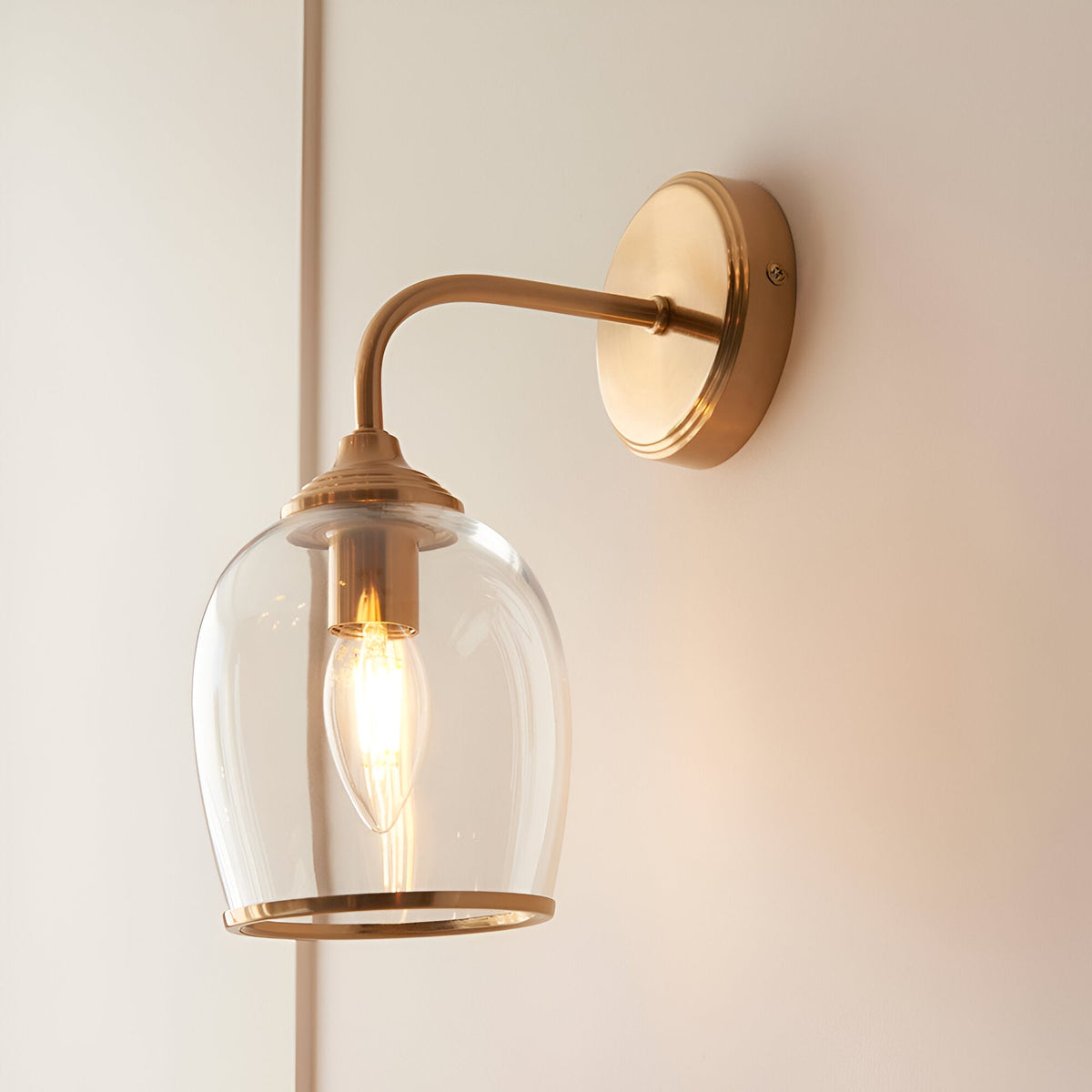 Halstead Wall Light in brushed brass, single arm with round back plate and clear domed glass shade, lit E14 bulb, mounted on a light wall.