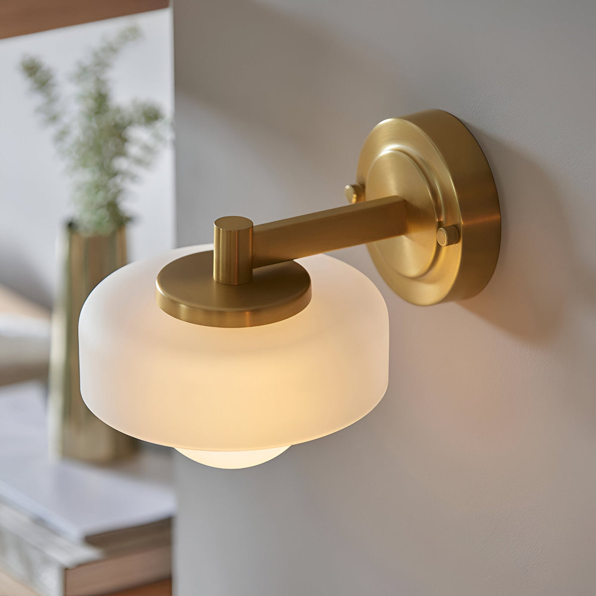 Endon Ashby brass wall light with frosted glass disc shade and opal sphere bulb in brushed aged brass, mounted beside a vase in a living space.