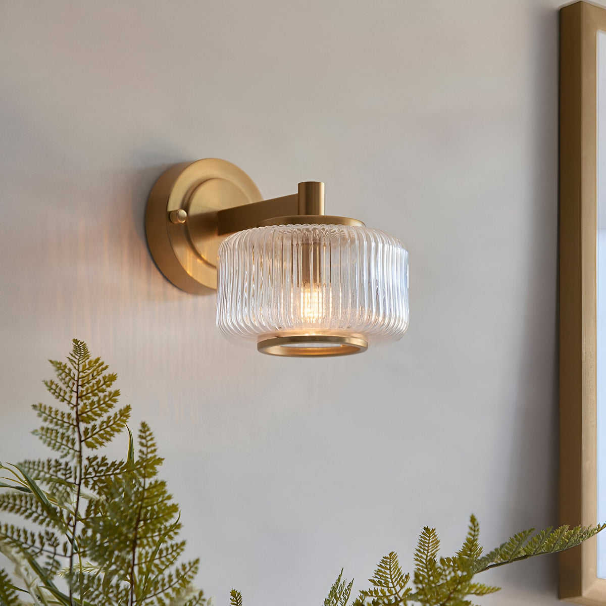 Endon Rutland wall light in brushed aged brass with a ribbed clear glass shade, mounted beside a mirror above ferns in a bedroom setting.