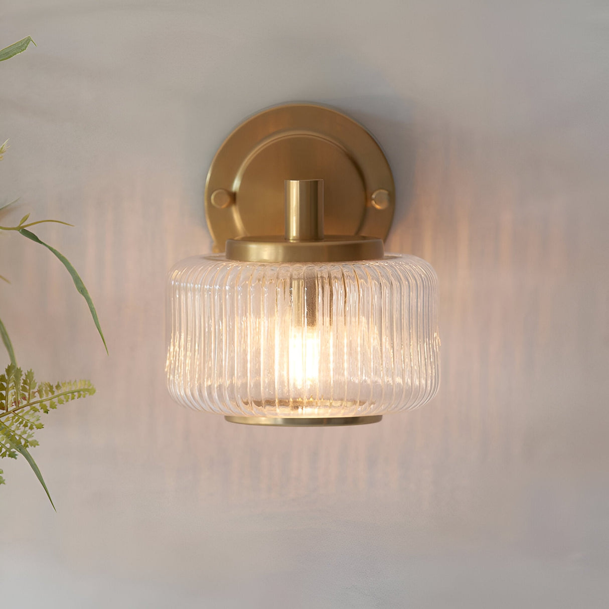 Endon Rutland wall light in brushed aged brass with a ribbed clear glass shade, illuminated and mounted on a neutral wall beside foliage.