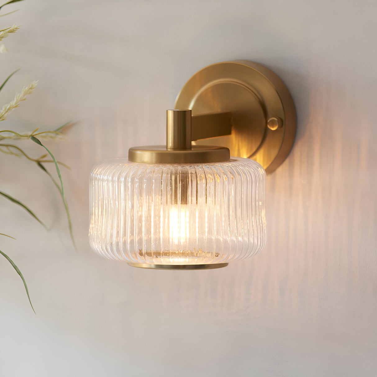 Endon Rutland wall light in brushed aged brass with a ribbed clear glass shade, illuminated on a light wall beside dried grasses.