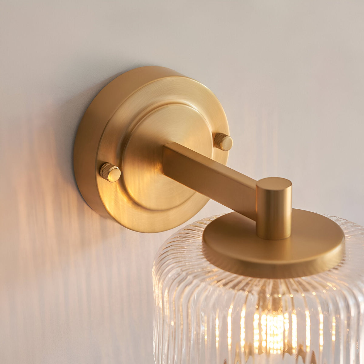Endon Rutland wall light close-up showing the brushed aged brass circular backplate, knurled screws, arm, and ribbed clear glass shade top.