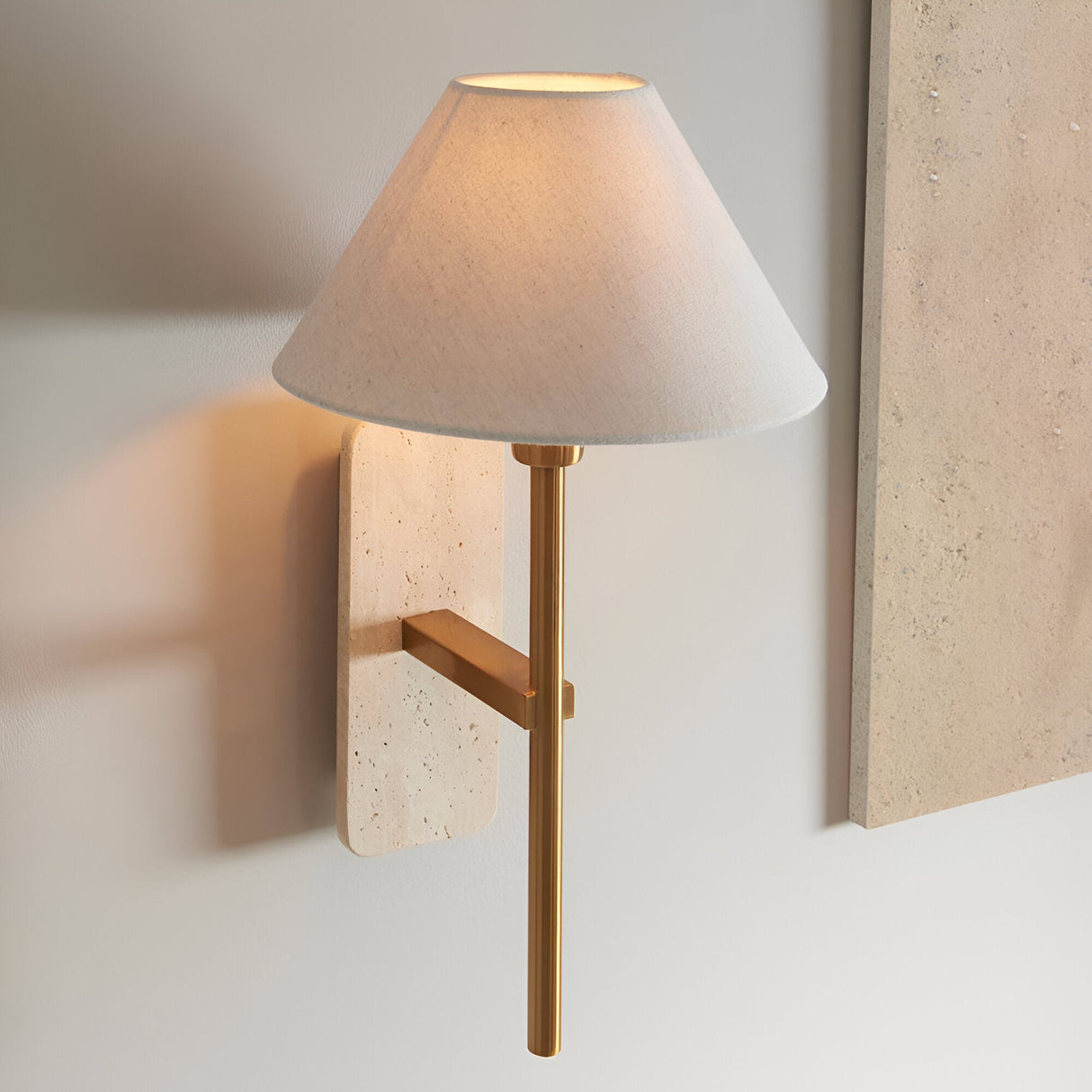 Bramble Wall Light in brass and natural stone, with tapered linen shade on a slim brass arm, mounted on a light wall beside a stone panel.