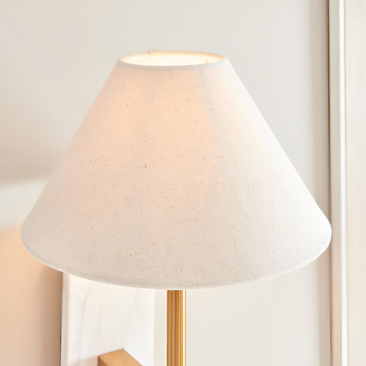 Bramble wall light with a tapered natural linen shade, brass-toned arm and marble backplate, shown lit against a plain light background.