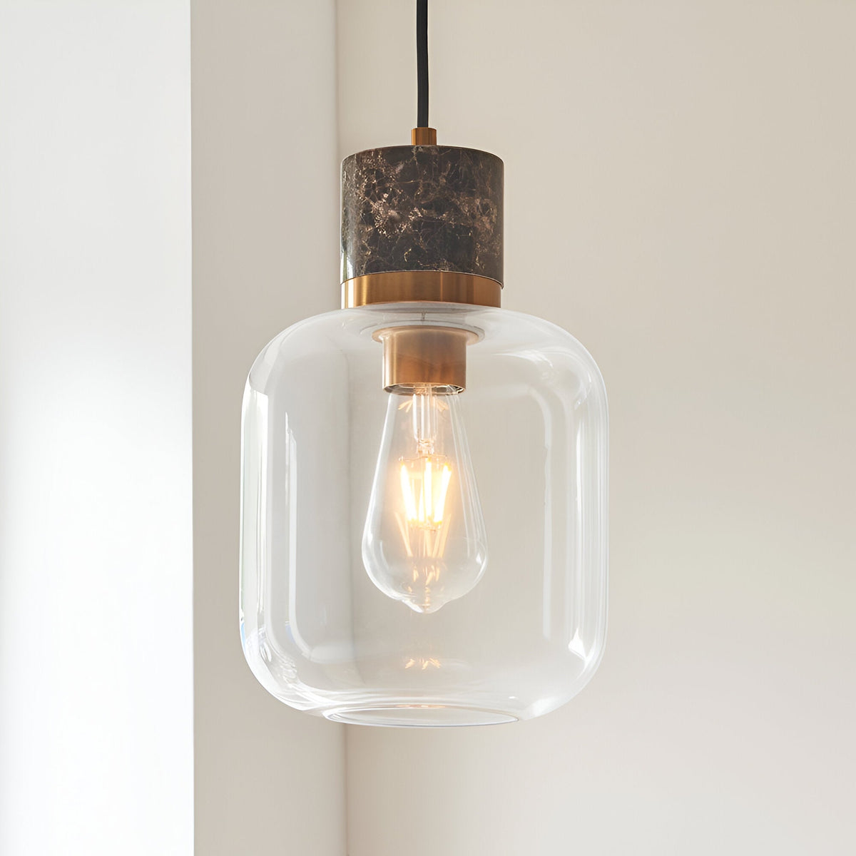Cranstead Pendant Light in brass and brown, with a marble-effect top, brass collar, and clear glass shade housing a glowing E27 bulb against a light wall.