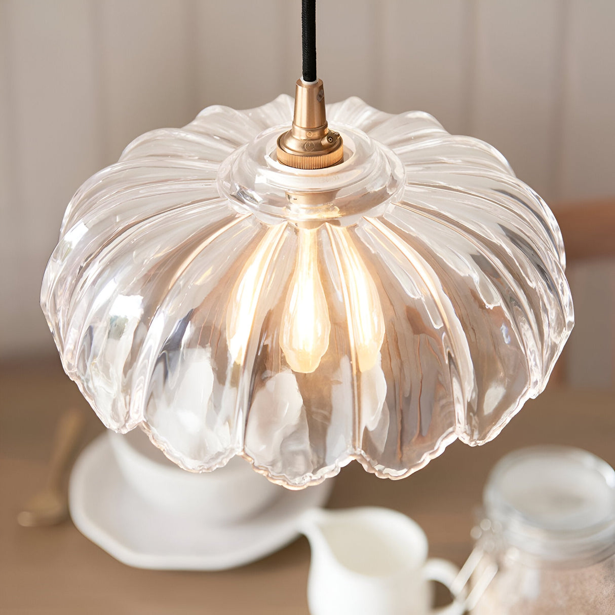 Brackmont Camellia pendant light with scalloped clear glass shade and brushed brass fitting on black cable, shown lit above a kitchen table.