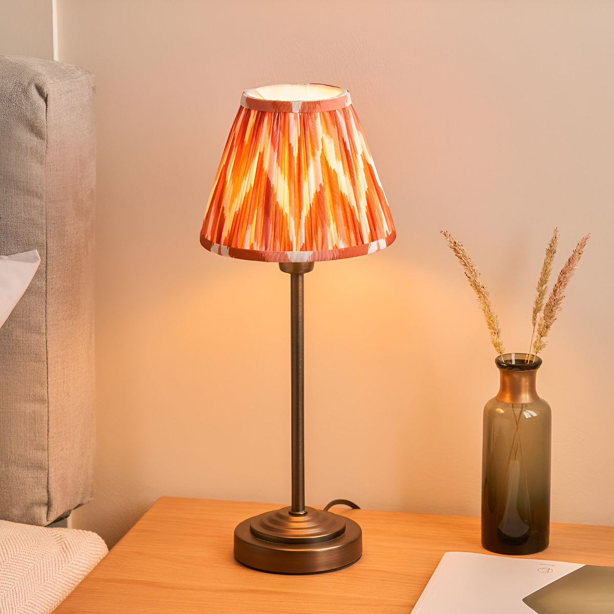 Endon Cliffbourne Zigzag table lamp with bronze stepped base and tapered pleated apricot orange zigzag shade, on a bedside table beside a glass vase.