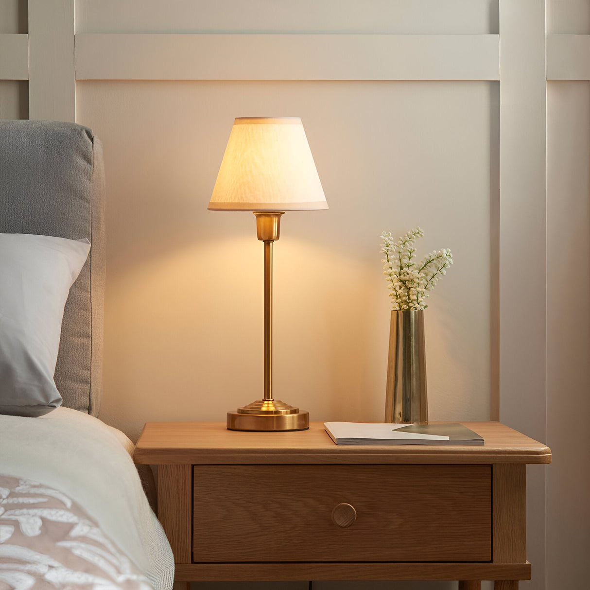 Cliffbourne Ivy Table Lamp in brass with a white tapered fabric shade, shown lit on a wooden bedside table beside a vase and headboard.