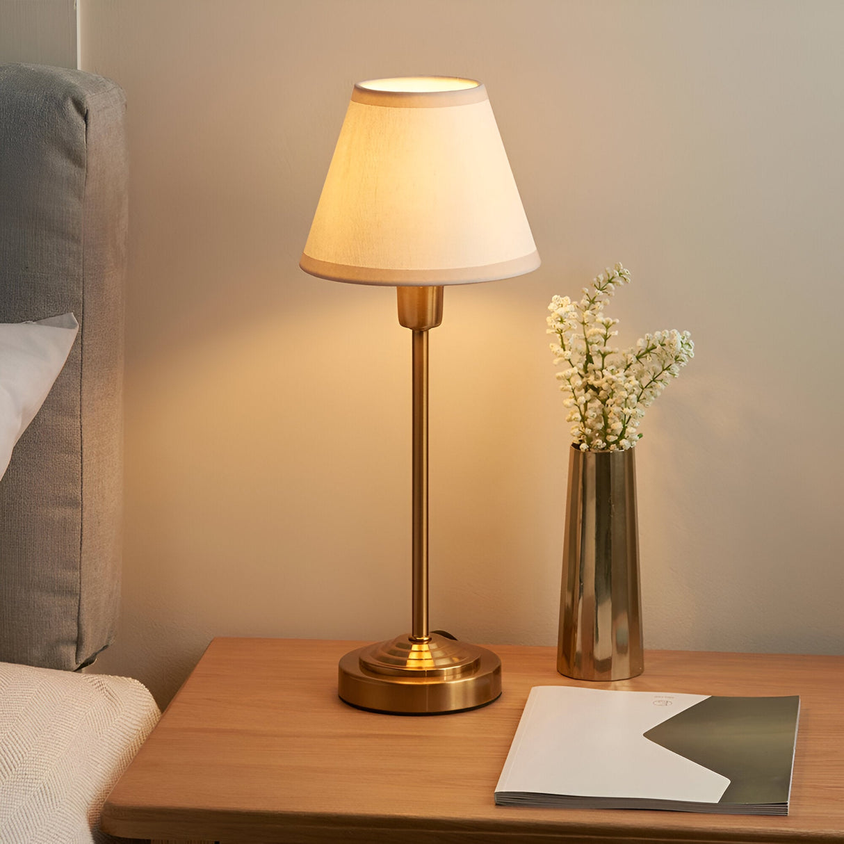 Cliffbourne Ivy Table Lamp with stepped brass base and tapered white fabric shade, lit on a wooden bedside table beside a vase of flowers.