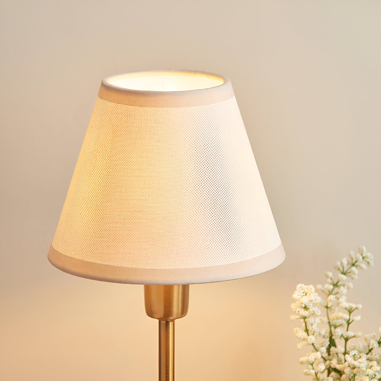 Cliffbourne Ivy Table Lamp with tapered white fabric shade and brass stepped metal base, shown lit against a neutral background with white flowers nearby.