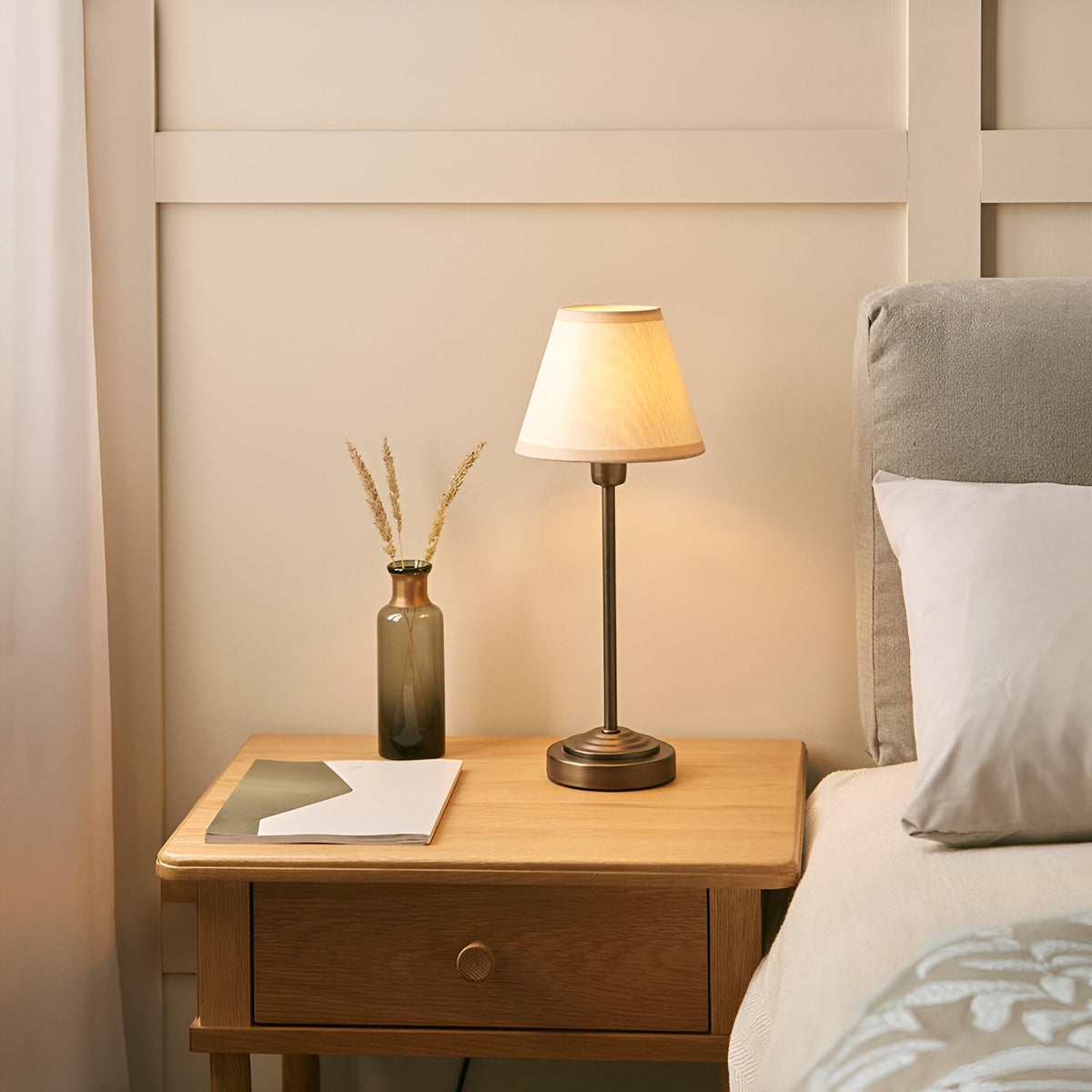 Endon Cliffbourne Ivy table lamp with stepped antique bronze base and tapered vintage white shade, lit on an oak bedside drawer beside a bed.