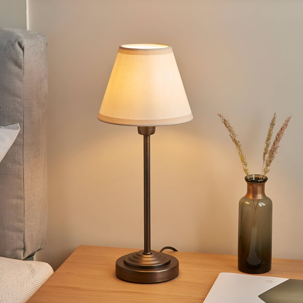 Endon Cliffbourne Ivy table lamp with stepped antique bronze base and tapered vintage white fabric shade, lit on a wooden bedside table beside a glass vase.