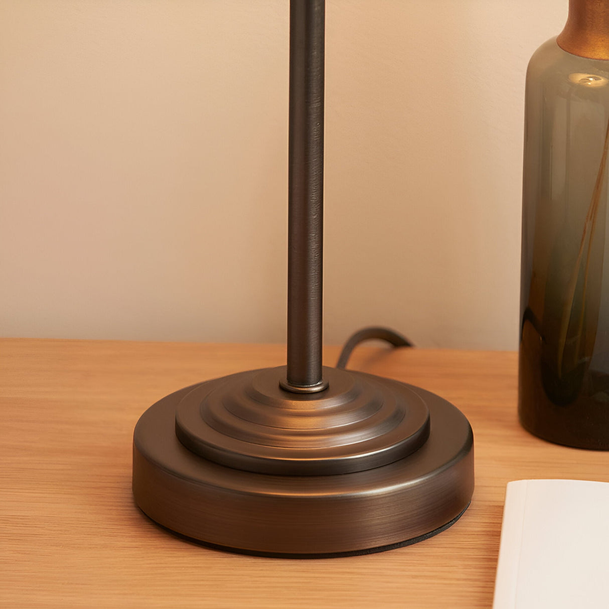 Close-up of the Endon Cliffbourne Ivy table lamp's stepped antique bronze base on a wooden surface beside a green glass vase.