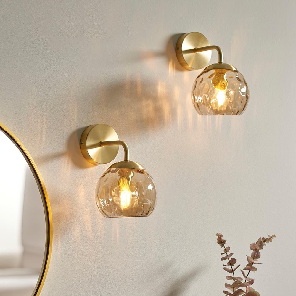 Endon Draycott wall lights in satin brass with champagne dimpled glass globe shades, mounted as a pair on a pale wall beside a round gold mirror.
