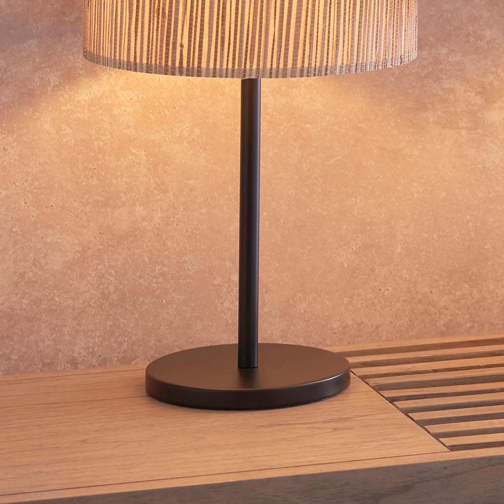 Salcombe Table Lamp in matt black metal with woven seagrass drum shade, shown lit on a wooden surface against a plaster wall.